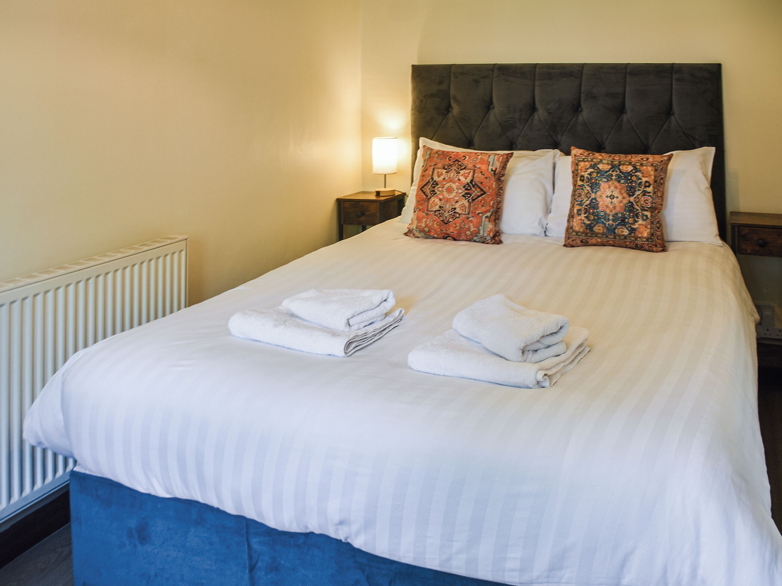 A neatly made bed with white linens and folded towels, a dark upholstered headboard, and decorative pillows with intricate patterns.