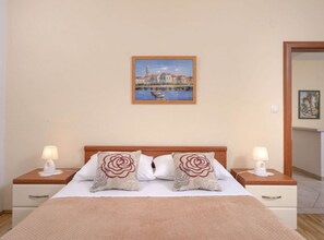 A hotel room with a bed, two bedside tables with lamps, a painting on the wall, and a door to another room. - One Bedroom Apartment With Balcony 3 (Jelsa)