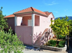 A pink building with a red-tiled roof, a staircase leading to a covered entrance, and a clear sky. - One Bedroom Apartment With Balcony 3 (Jelsa)