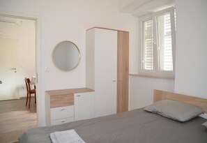 Ein Schlafzimmer mit Bett, Nachttisch, Kleiderschrank, rundem Spiegel und einem Fenster mit Rollläden.