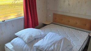 A single bed with a headboard, a window with a red curtain, and a wall with a floral pattern. - Country Bungalow (Saint-Georges-de-Reintembault)