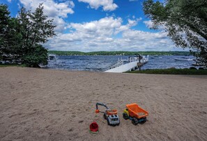 Ein sandiger Strand mit einem Steg, der in einen See führt, umgeben von Bäumen und Booten.