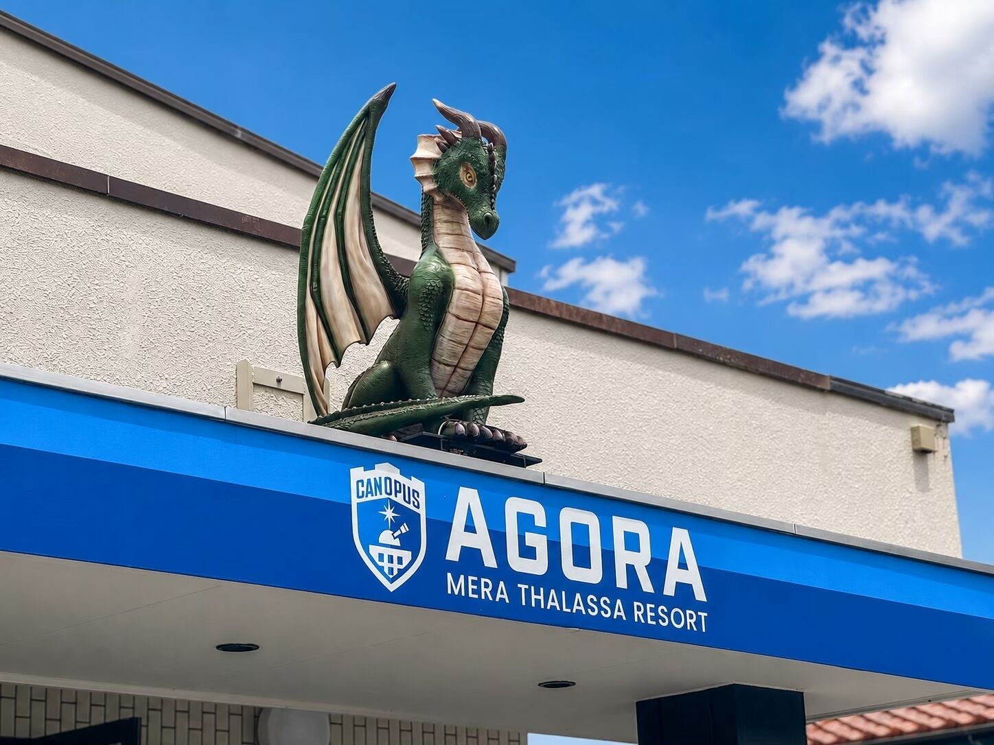 Een drakenbeeld op een gebouw met een blauw en wit bord waarop staat: 'AGORA MERA THALASSA RESORT'.