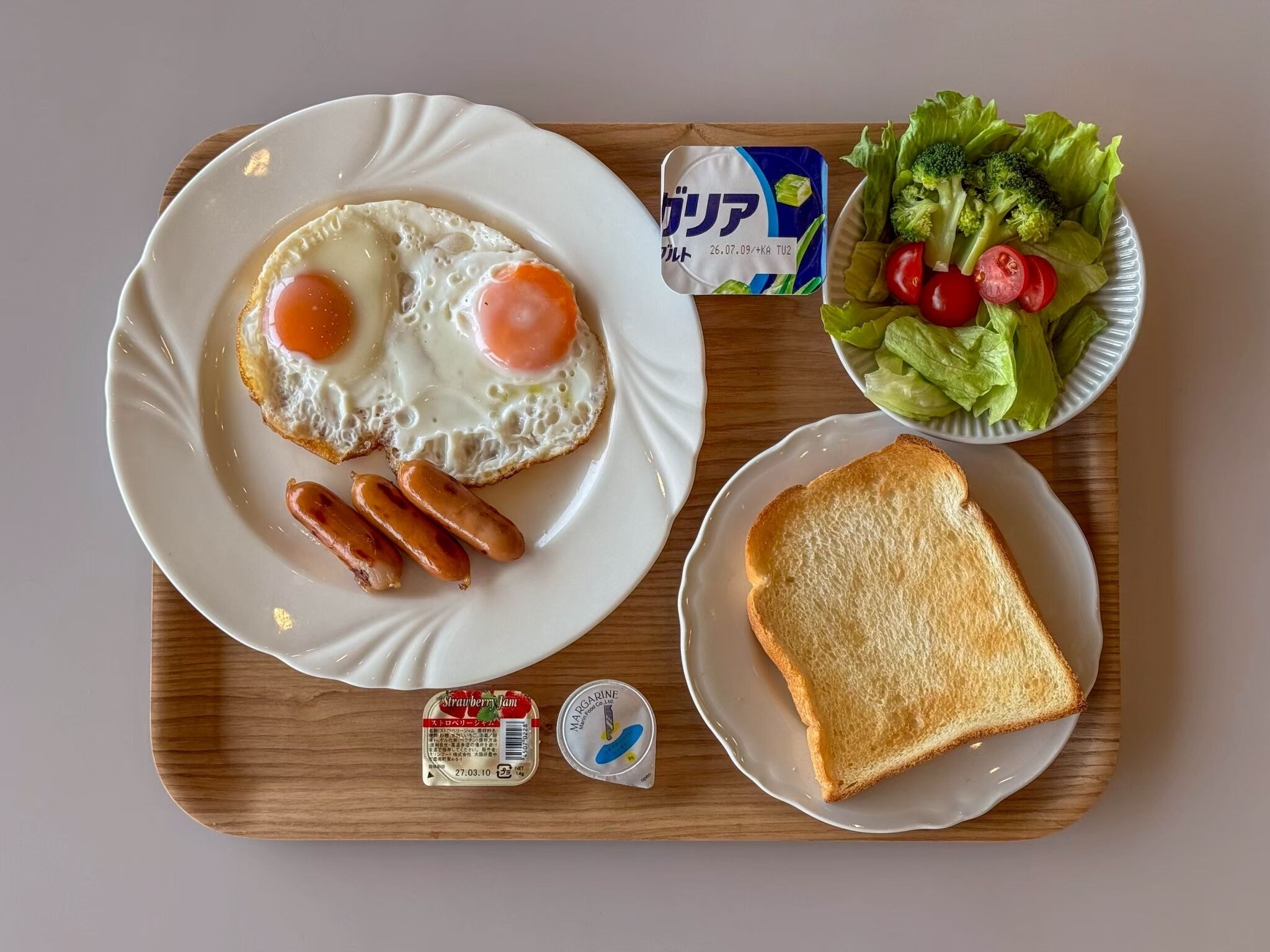 A meal tray with a plate of fried eggs, sausages, a bowl of salad, a slice of toast, and condiments.