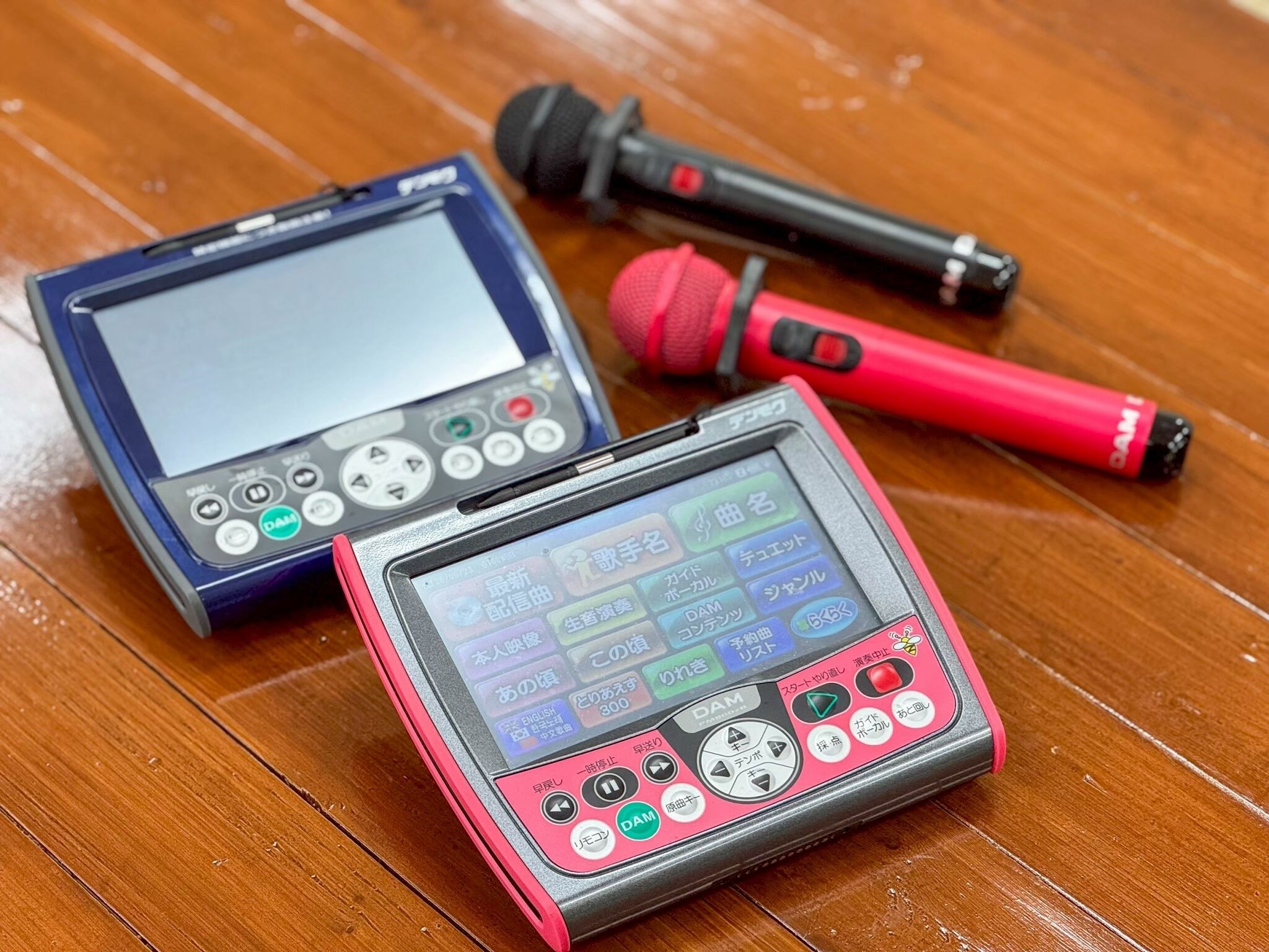 Un dispositivo electrónico portátil con pantalla y botones, un micrófono rojo y un micrófono negro sobre una superficie de madera.