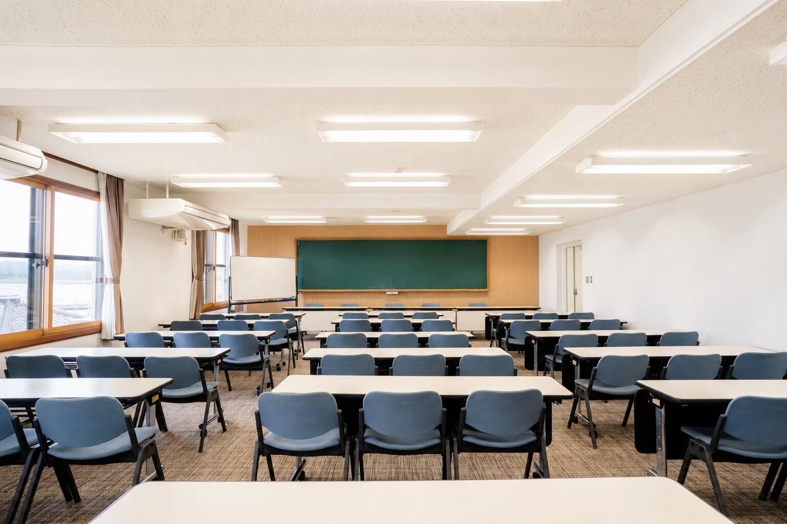 A classroom with rows of tables and chairs, a large green chalkboard, and a whiteboard.