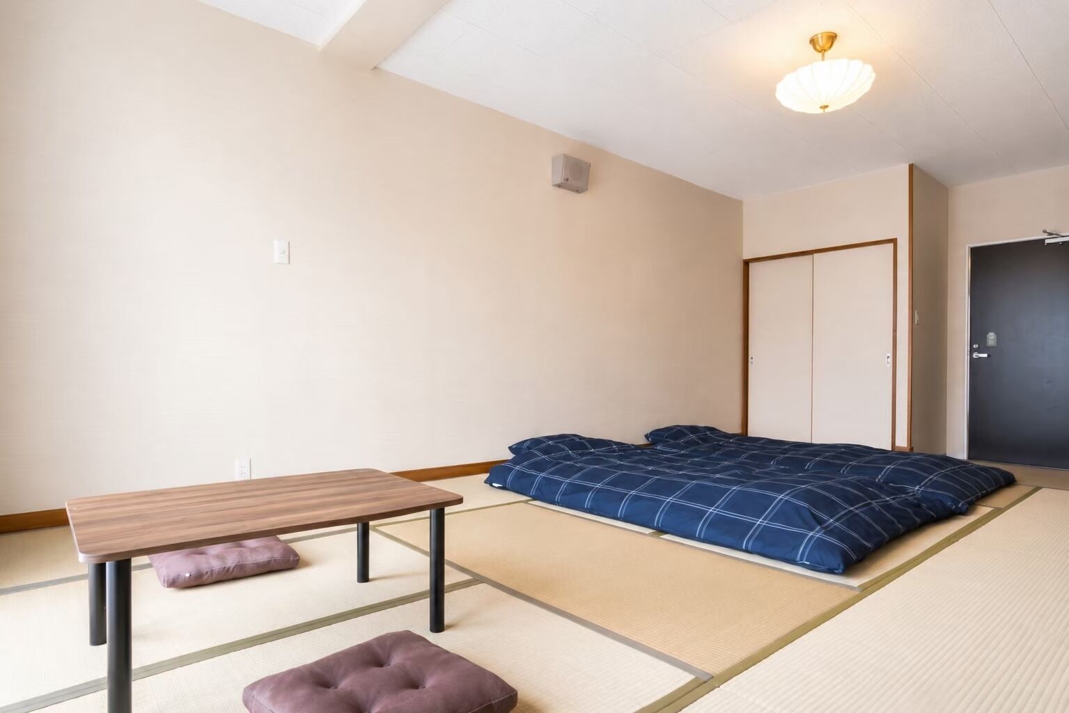 Ein Zimmer mit Tatami-Boden, einem niedrigen Holztisch, einem Bett mit blauer Bettwäsche und einer dunklen Tür.