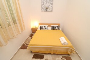 A bedroom with a bed, pillows, a nightstand, a lamp, a window with curtains, and a wall art piece. - Apartments K & N -one Bedroom Apartment (Blato)