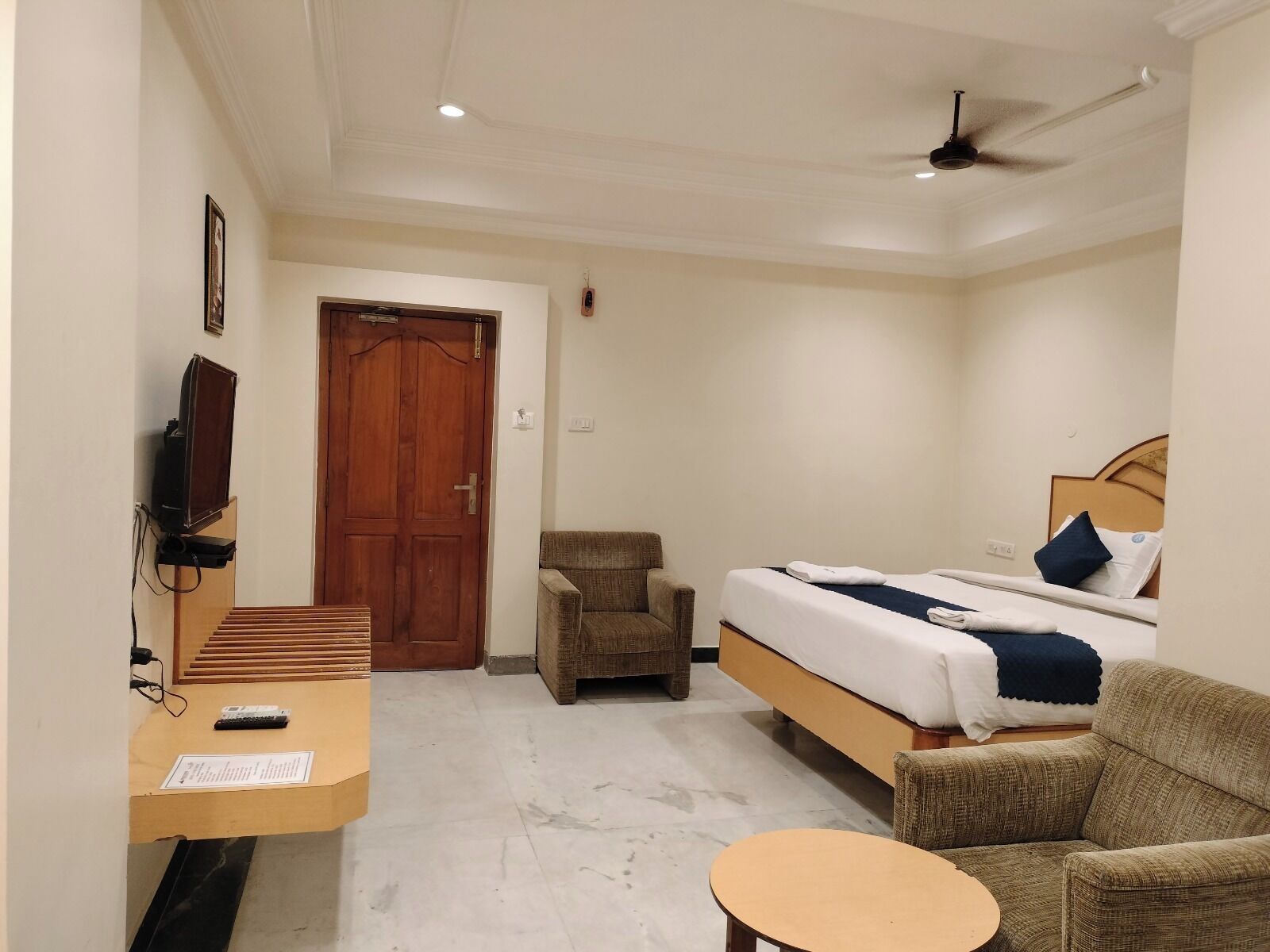 Habitación de hotel con cama, una silla, una mesita, televisor y ventilador de techo.