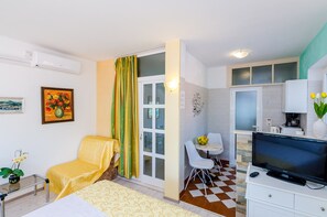 Ein gemütlicher, kompakter Raum mit Bett, Stuhl, kleinem Tisch, Fernseher und Küchenzeile.