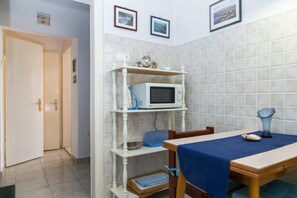 Ein Zimmer mit Fliesenwand, einer auf einem Regal stehenden Mikrowelle, einem Tisch mit blauer Tischdecke und einer Tür zu einem anderen Raum.