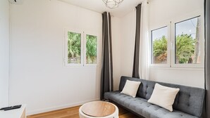 Uma sala de estar moderna com um sofá cinzento, duas janelas com cortinas, chão de madeira e um tapete redondo.