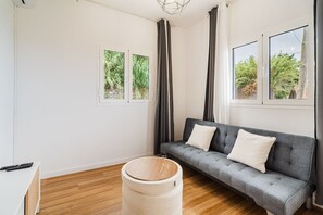 Ein modernes Wohnzimmer mit einem grauen Sofa, zwei mit Vorhängen versehenen Fenstern, einem Holzboden und einem runden Teppich.
