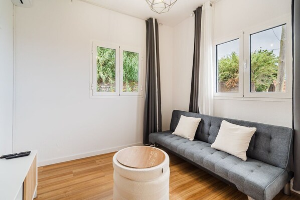 Ein modernes Wohnzimmer mit einem grauen Sofa, zwei mit Vorhängen versehenen Fenstern, einem Holzboden und einem runden Teppich.