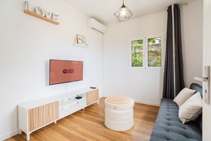 A modern living room with a flat-screen TV, a white console table, a grey sofa, a round ottoman, and a window with a view of greenery. - GuestReady - Charming Retreat with Patio & Views (Porto Moniz)