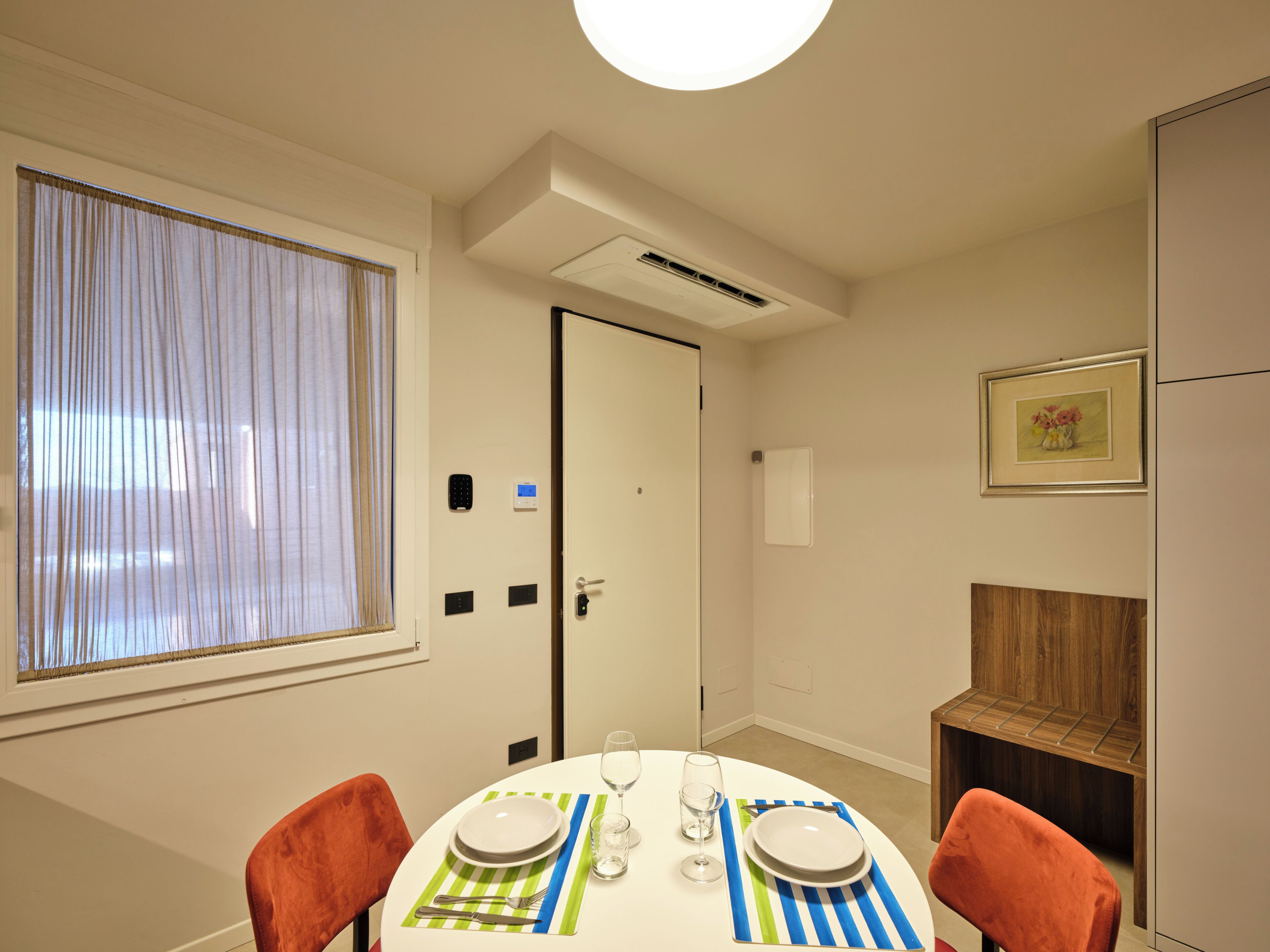 A dining area with a round table set for two, a window with curtains, an air conditioning unit, and a door with a lock.