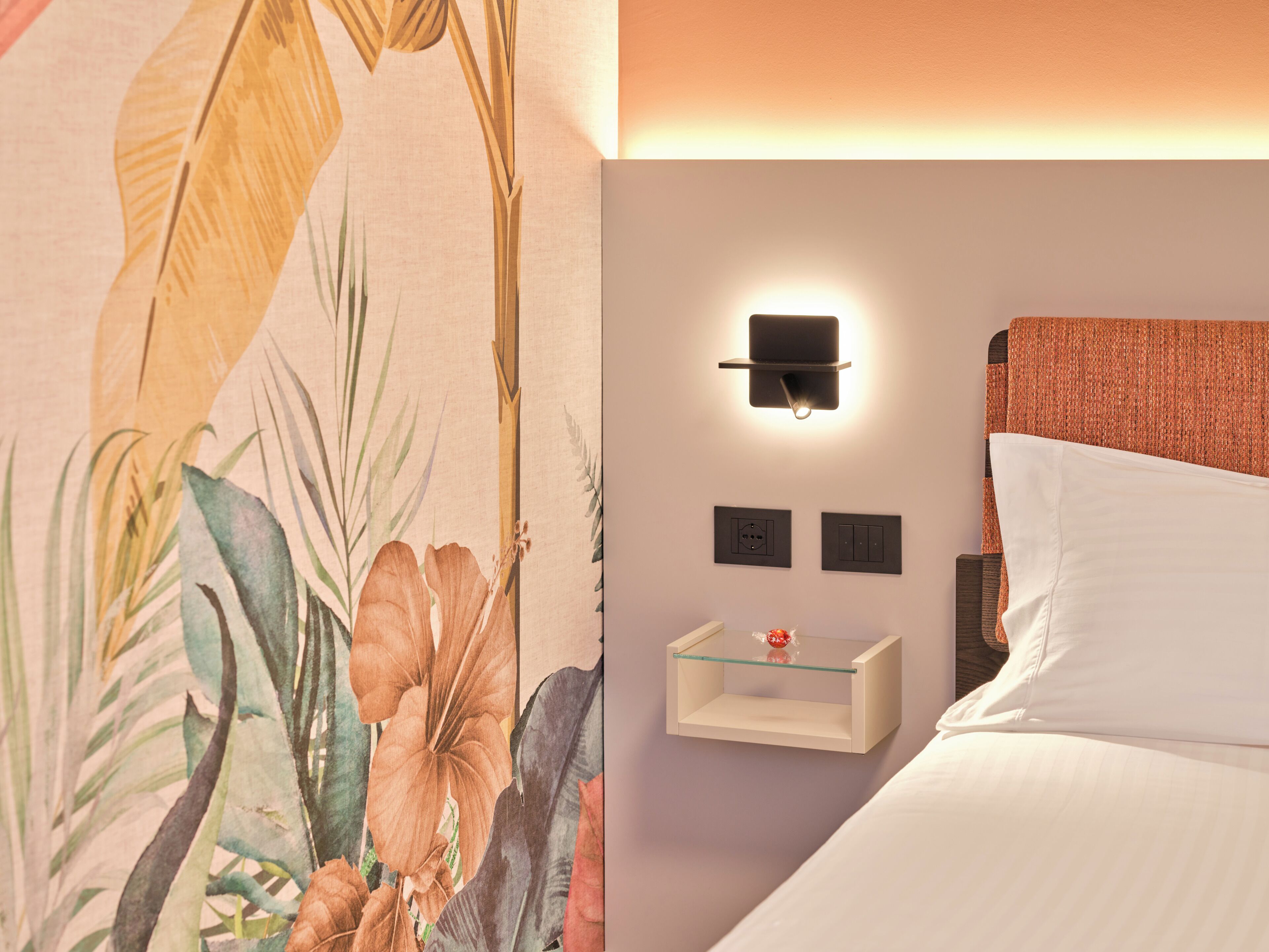 Una habitación de hotel con cama, mesita de noche, lámpara de pared y toma de corriente.