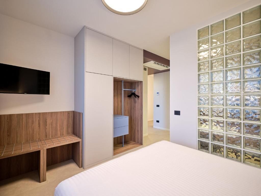 Um quarto com cama, guarda-roupa, um banco, televisão e uma parede de vidro.