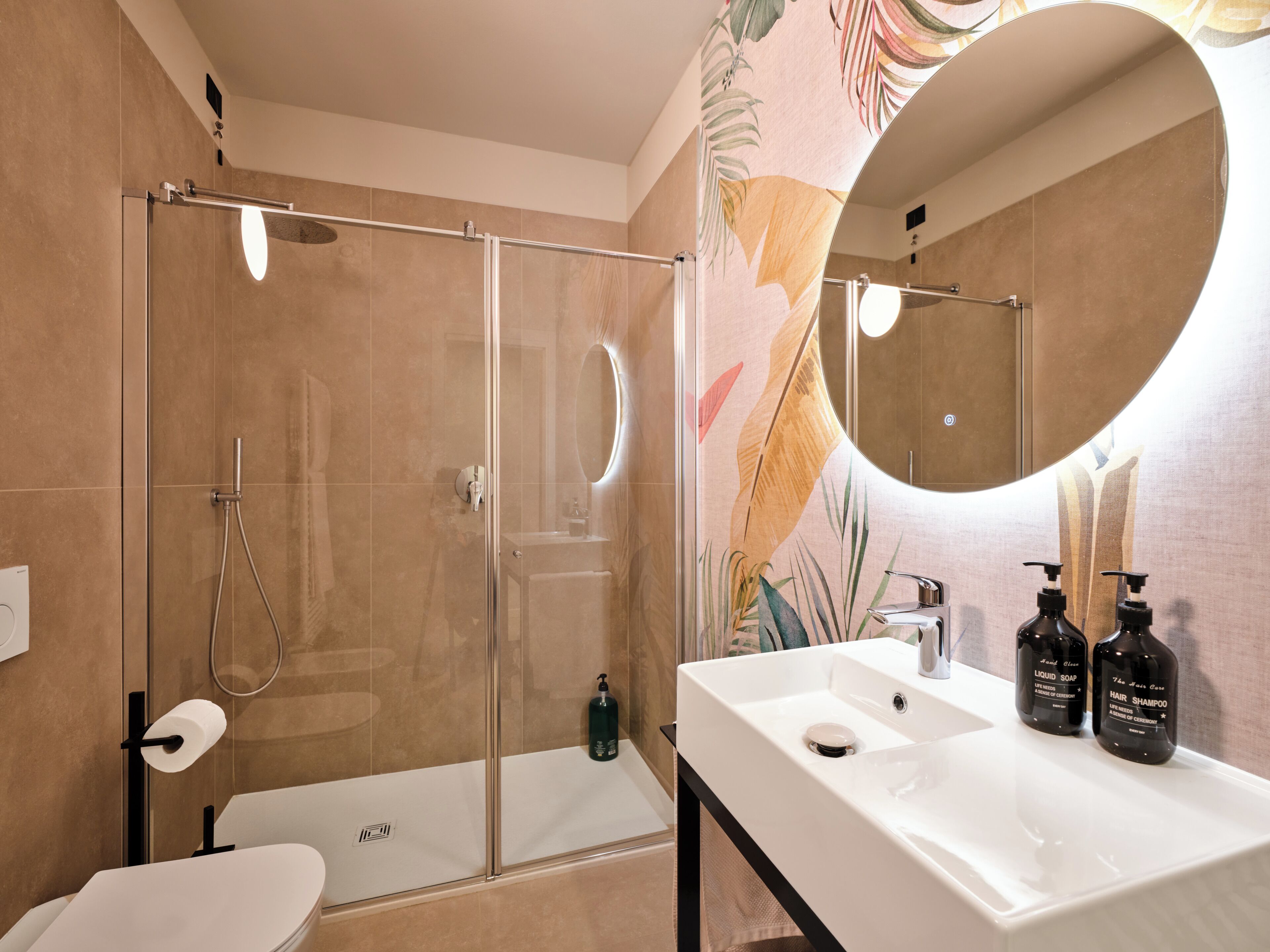 Une salle de bain moderne avec une douche à parois de verre, un lavabo blanc avec deux bouteilles de shampoing et un grand miroir.