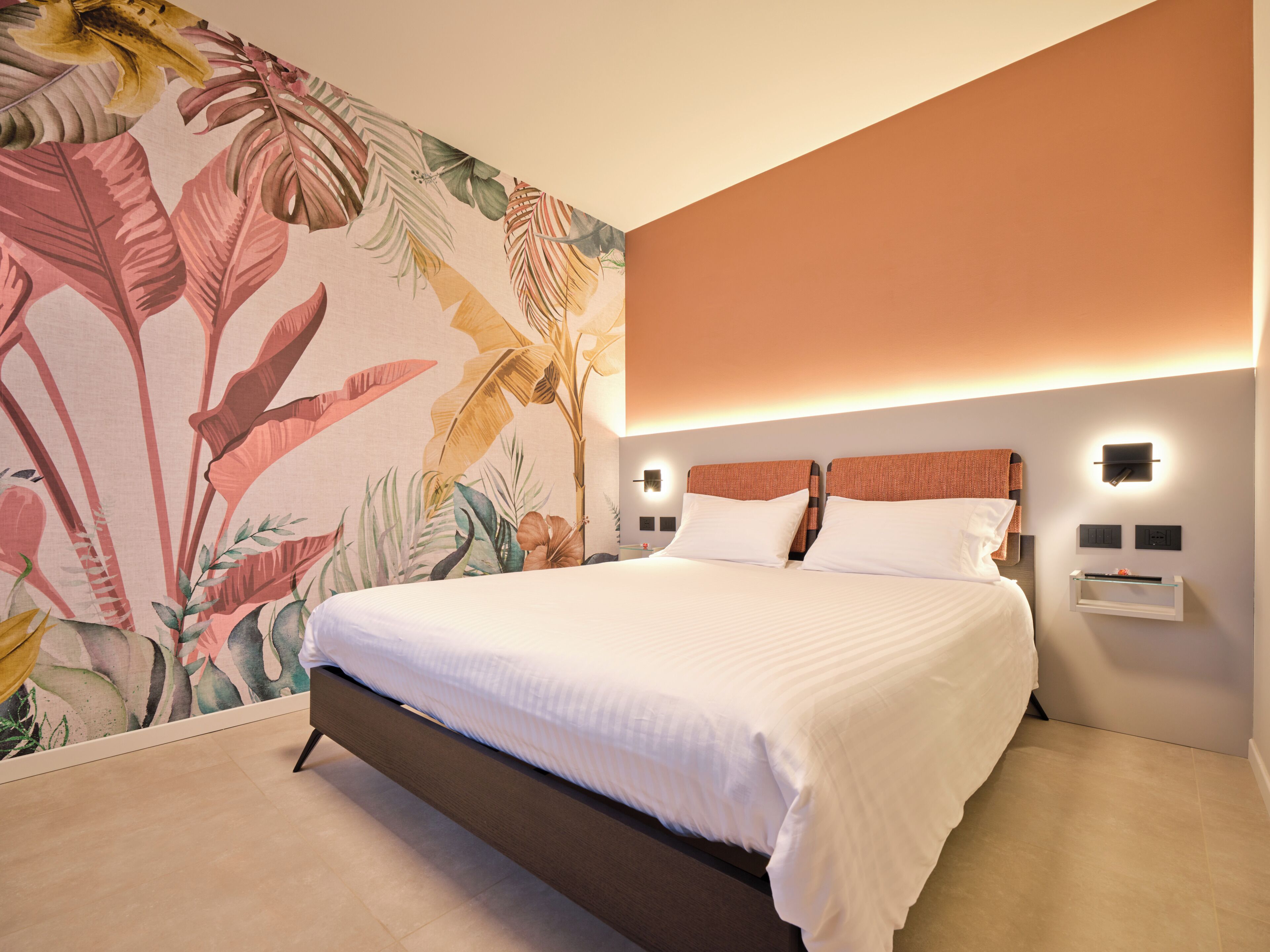 A hotel room with a bed, a nightstand, a wall-mounted light, and a tropical-themed wallpaper.