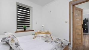 Una camera da letto con un letto, una finestra con persiane, un comodino con una tazza e un piattino, e una porta in legno.