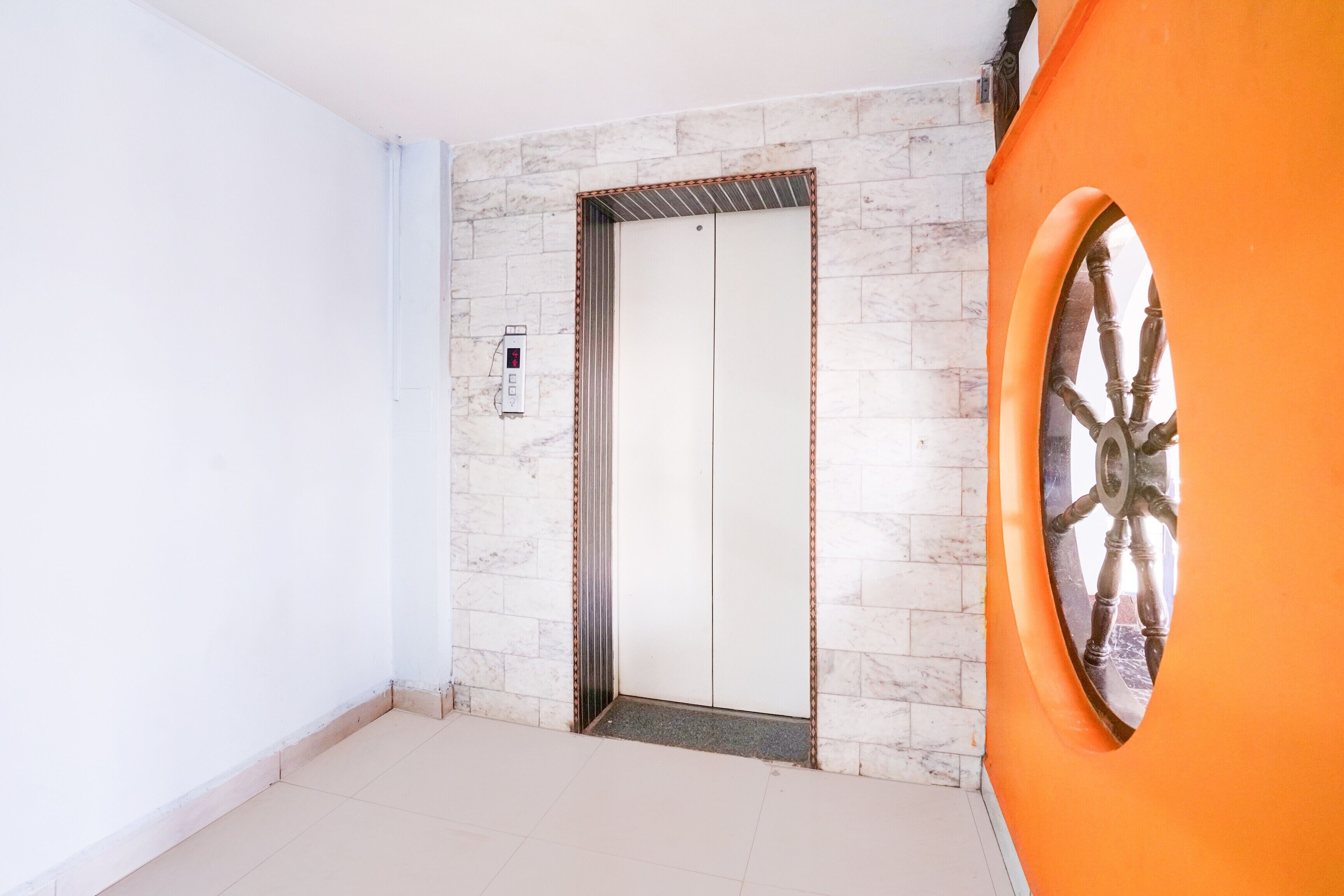 Una habitación con piso de baldosas blancas, pared de mármol, un ascensor y una puerta anaranjada grande con un detalle decorativo en forma de rueda.