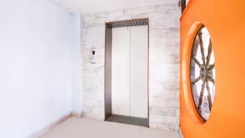 Una habitación con piso de baldosas blancas, pared de mármol, un ascensor y una puerta anaranjada grande con un detalle decorativo en forma de rueda.