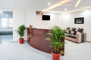 A reception area with a wooden reception desk, potted plants, a flat-screen TV, and a couch with patterned cushions. - Collection O Peenya 2nd stage (Bengaluru)