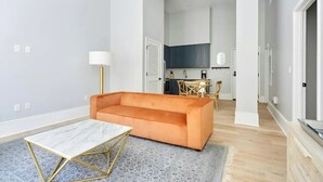 Uma sala de estar moderna com sofá, mesa de centro e abajur. Cozinha ao fundo, com armários e bancada.