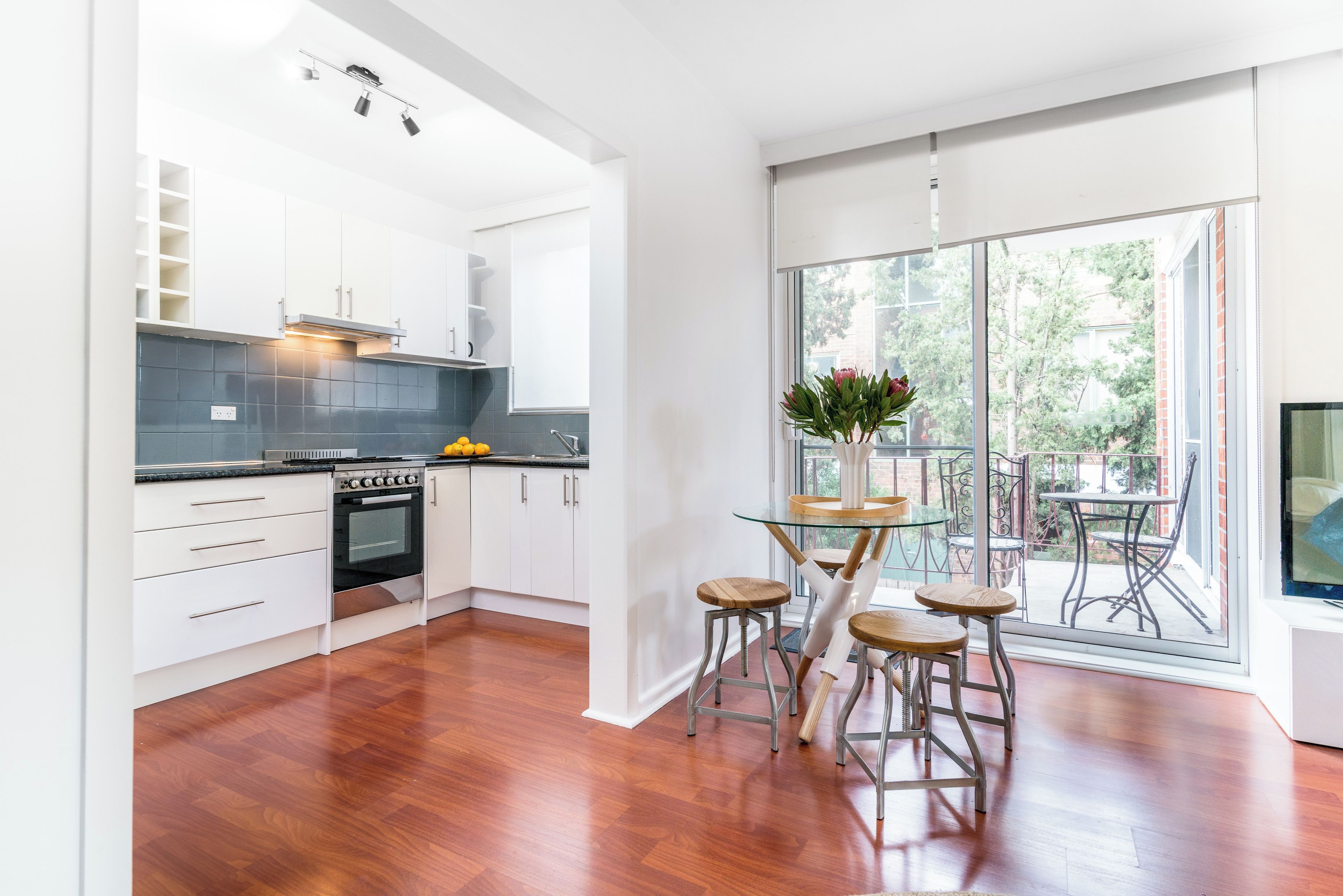 A modern kitchen with white cabinets, a built-in oven, and a dining area with a glass table and metal stools.