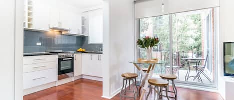 A modern kitchen with white cabinets, a built-in oven, and a dining area with a glass table and metal stools.