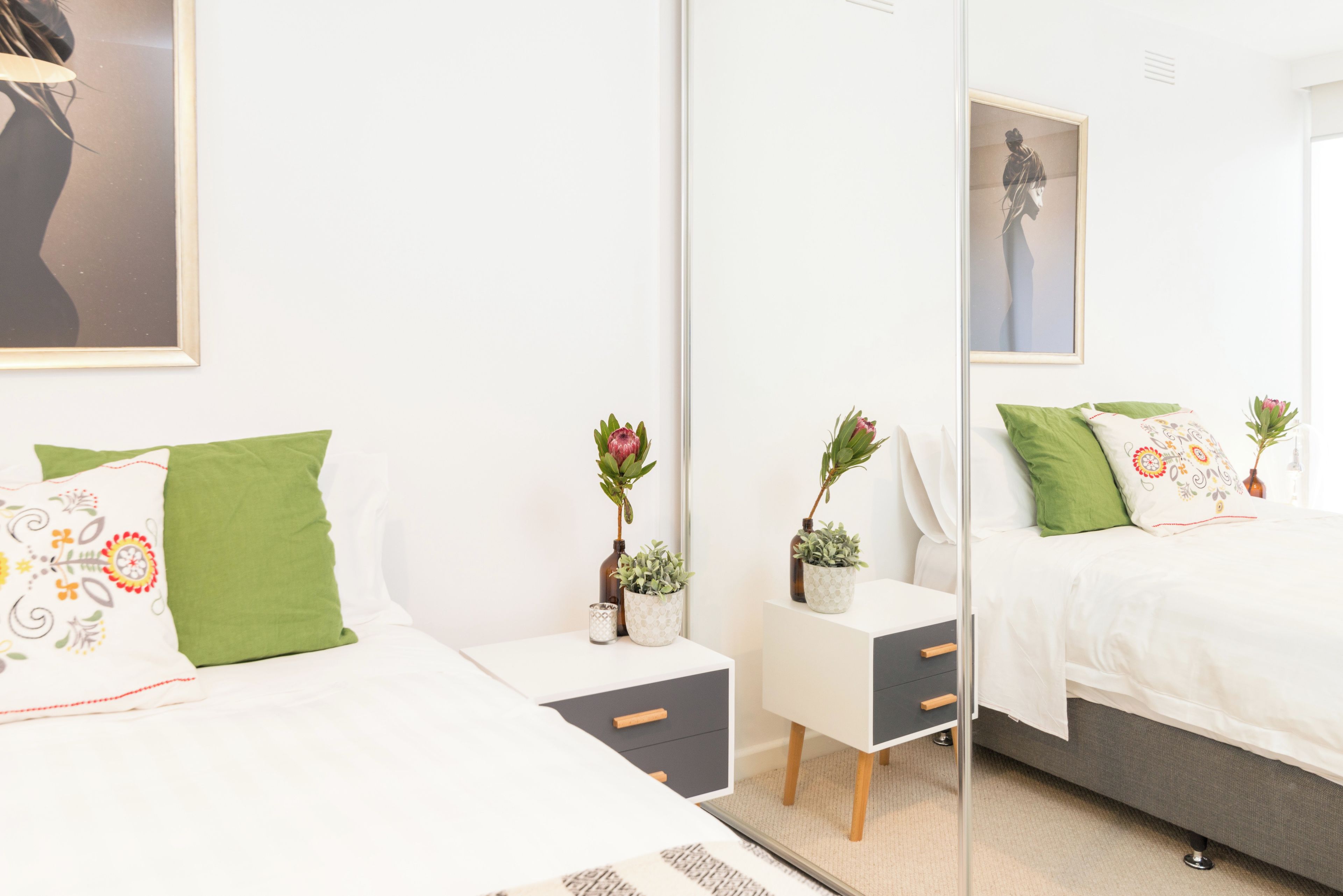 Et moderne soveværelse med et stort spejl, to senge med grønne puder, et lille bord med en plante og et natbord med en lampe.