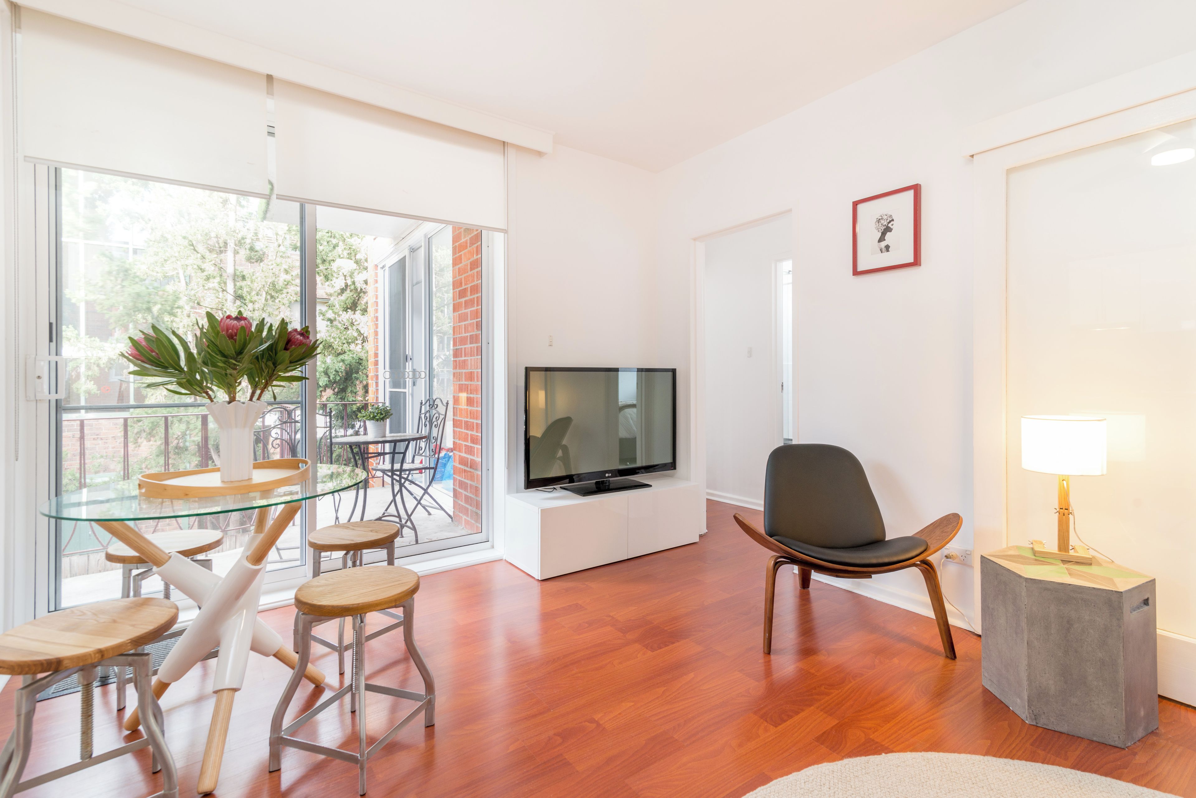 A modern living room with a glass dining table, a flat-screen TV, a comfortable chair, and a small side table with a lamp.