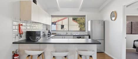 A modern kitchen with a central island, bar stools, a fire extinguisher, and a window with a view of the outside.