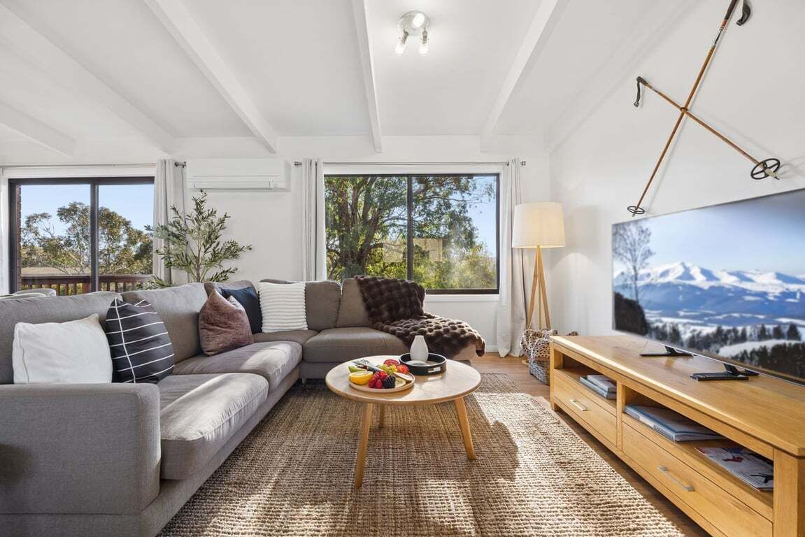 A modern living room with a grey sofa, a round coffee table with fruit, a wooden TV stand, and a large window with a mountain view.