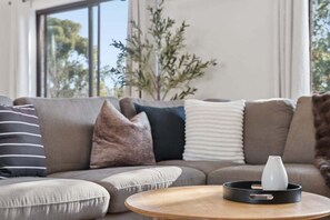 Ein modernes Wohnzimmer mit einem grauen Sofa, mehreren Kissen, einem runden Holztisch und einer Vase auf einem Tablett.