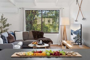 Ein modernes Wohnzimmer mit einer Couch, einem Couchtisch mit Obst, einem Gemälde und einem Fenster mit Blick auf Bäume.
