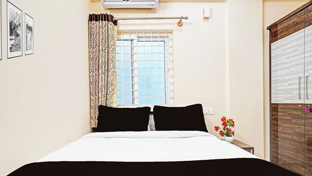 A bedroom with a bed, a window with a view, a curtain, a wall-mounted air conditioner, and a small table with a vase of flowers. - Collection O KR Puram Ayyappa Nagar (Bengaluru)