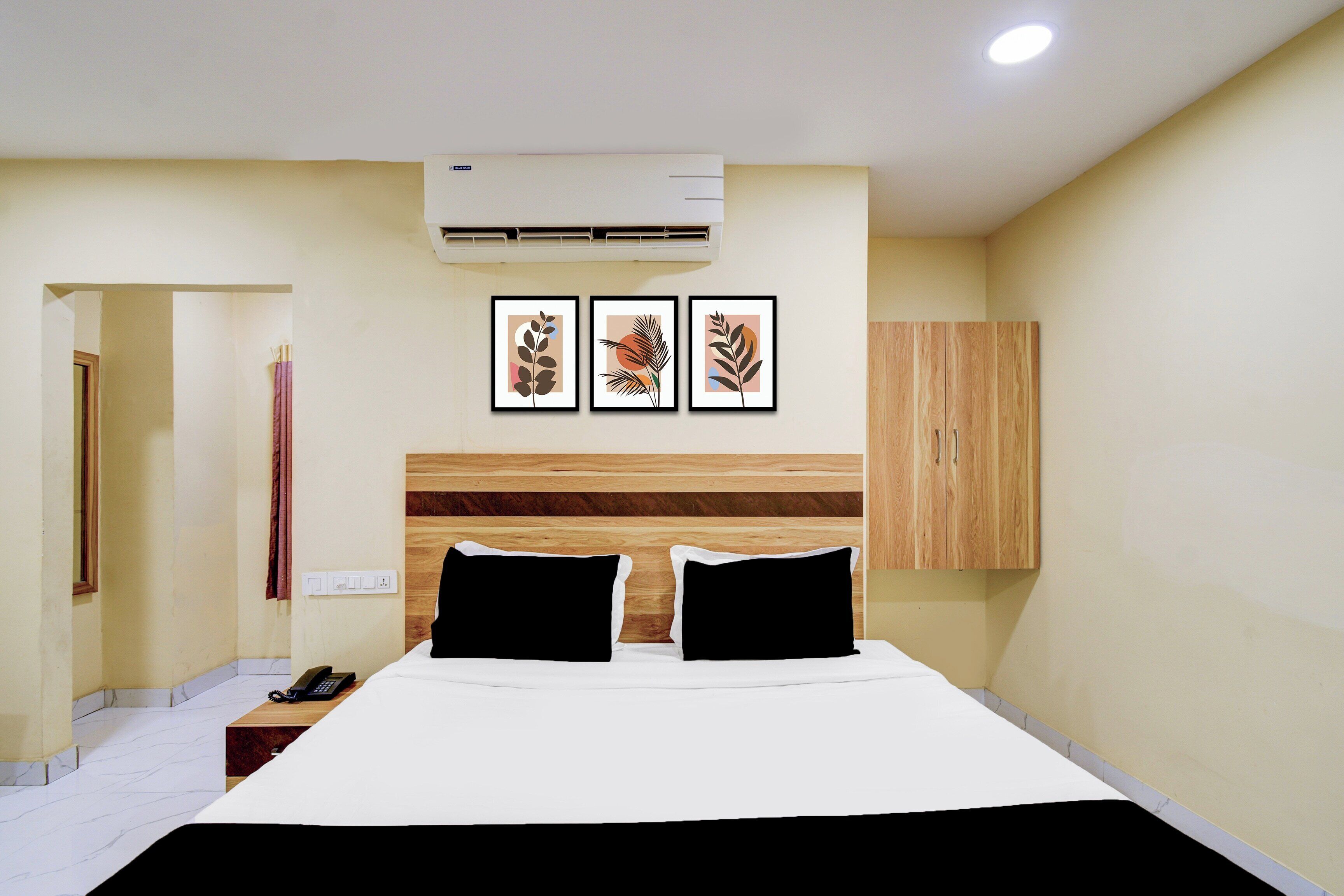 A hotel room with a bed, a wooden headboard, an air conditioning unit, and three framed artworks on the wall.