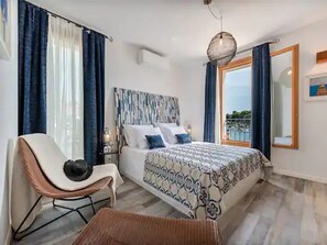 Ein Schlafzimmer mit Bett, einem Sessel, einem Nachttisch, einer Lampe und Blick ins Freie.