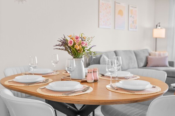 Mesa redonda de comedor dispuesta con platos, cubiertos y vasos, rodeada de sillas blancas.