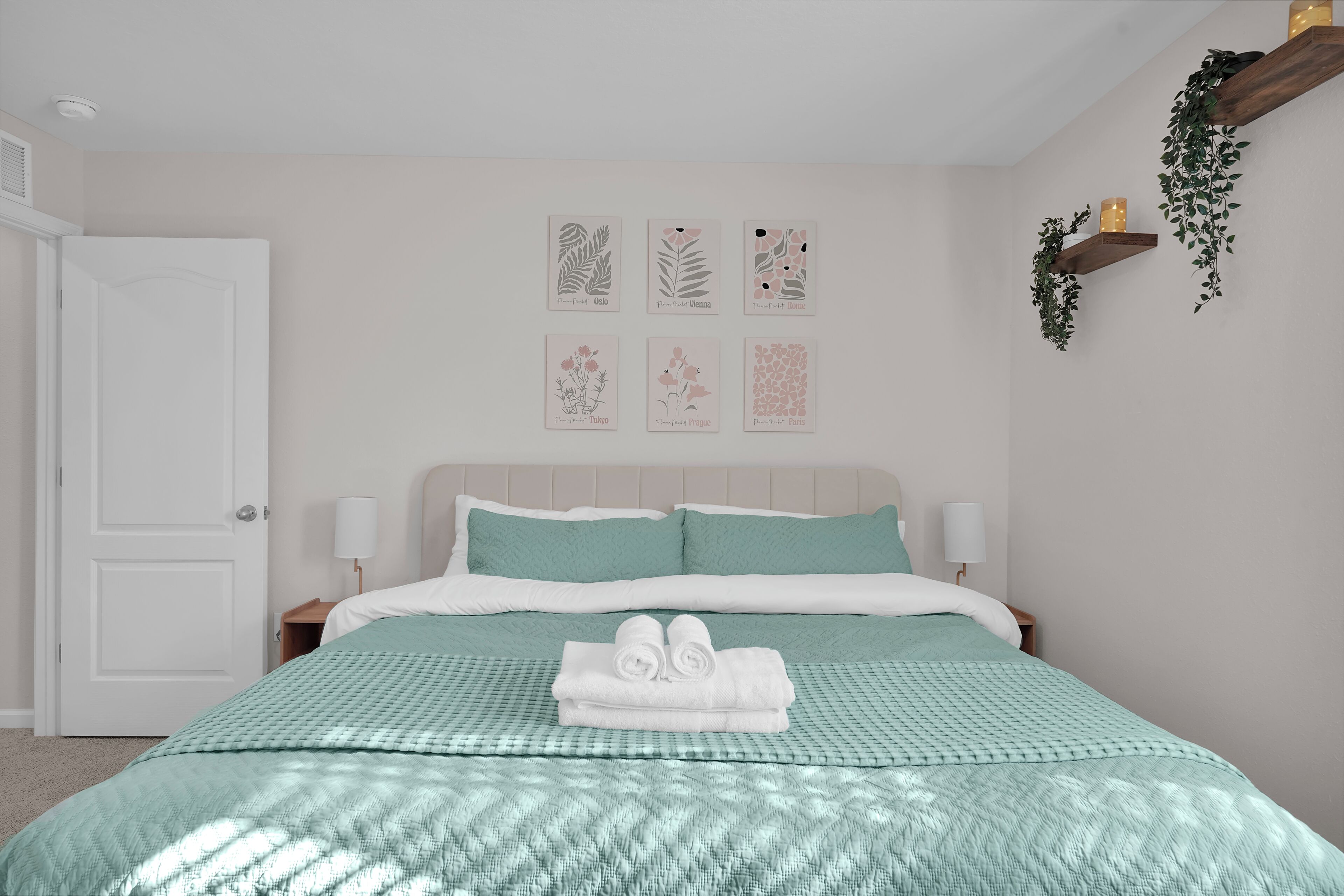 Un lit bien fait, recouvert d’une couvre-lit turquoise et agrémenté d’oreillers blancs, une porte blanche et des œuvres d’art murales représentant des fleurs provenant de différentes villes.