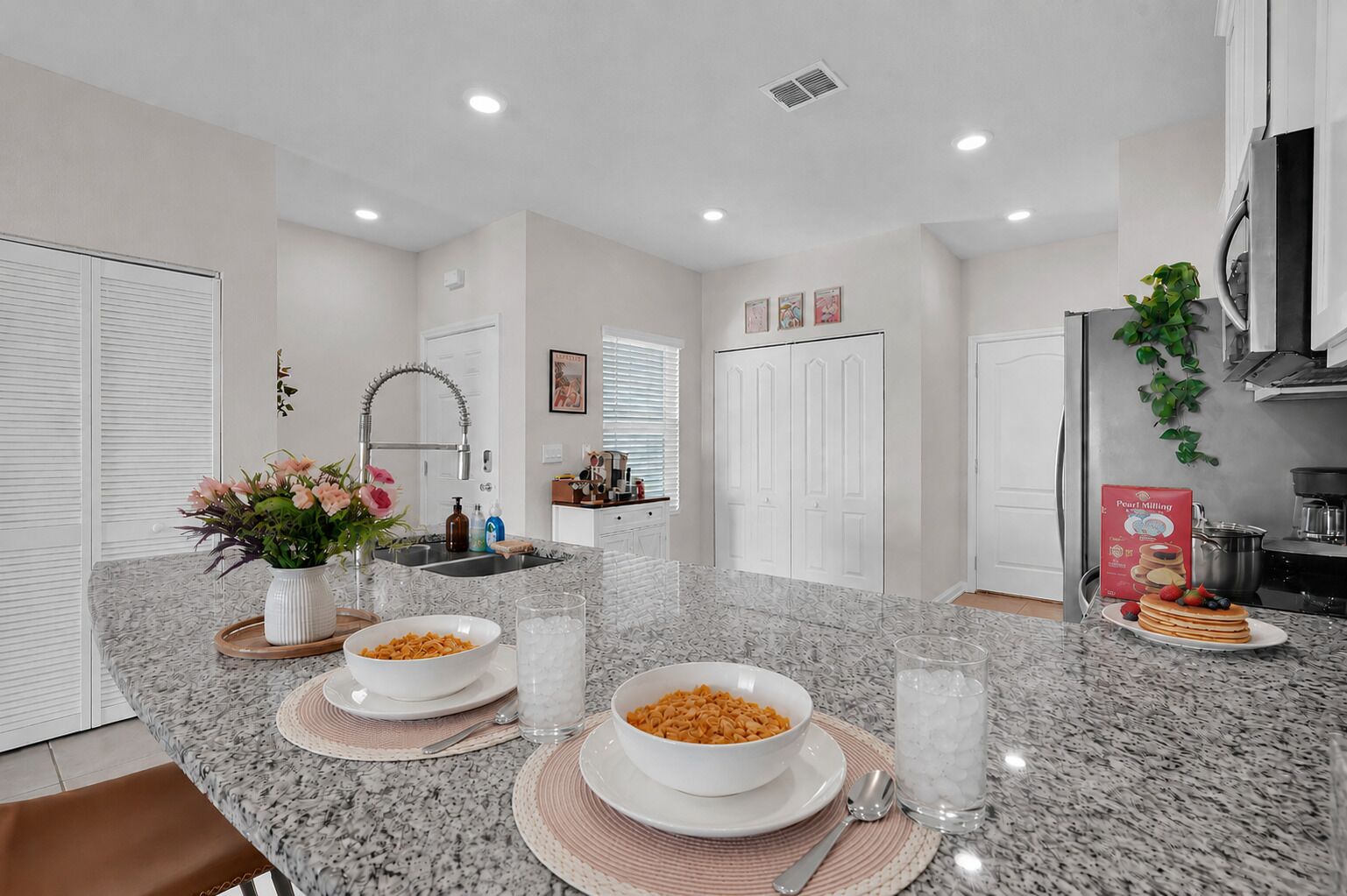 A modern kitchen with granite countertops, white cabinetry, and a breakfast setup with cereal bowls and a plate of pancakes.
