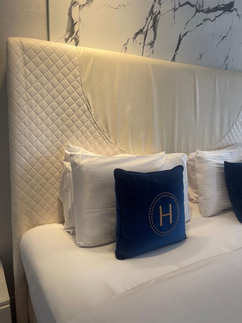 A bed with a quilted headboard, white bedding, and a dark blue pillow with a gold letter 'H'.