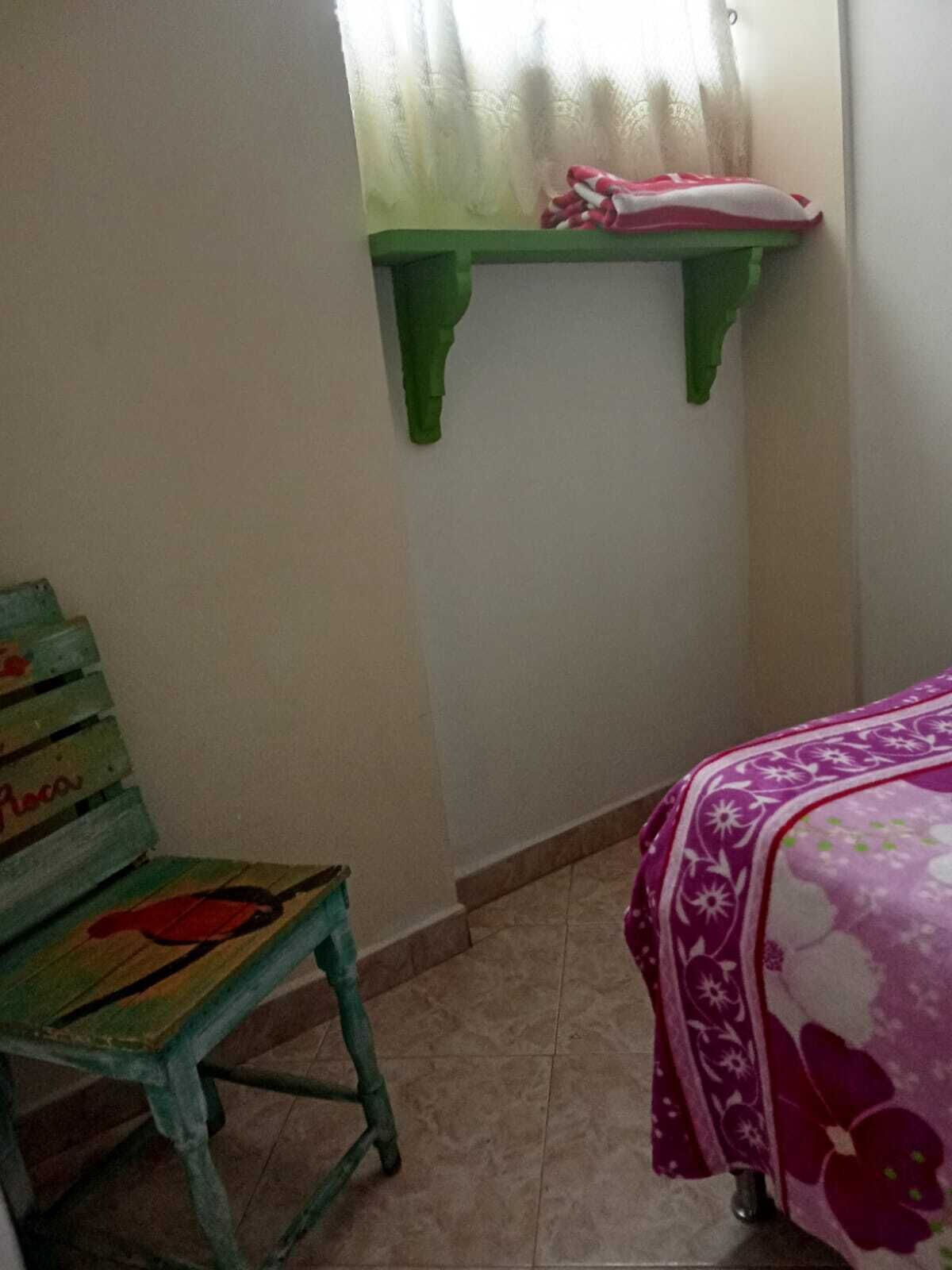Une pièce avec une chaise verte, un lit recouvert d’une courtepointe à motifs floraux violets et une étagère sur laquelle se trouve un objet rose.