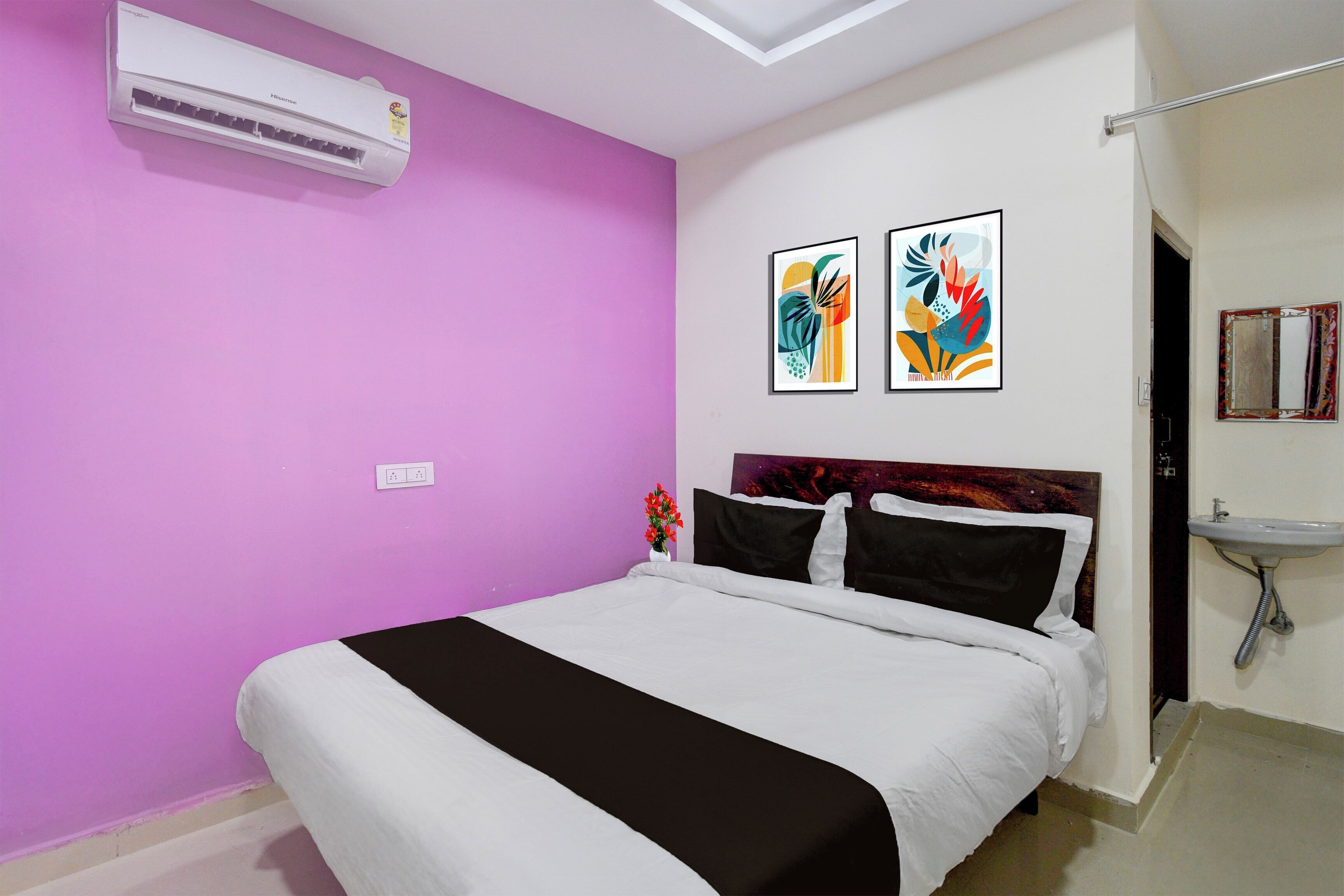 A bedroom with a bed, a wall-mounted air conditioner, a bedside table with a vase of flowers, and two framed artworks on the wall.