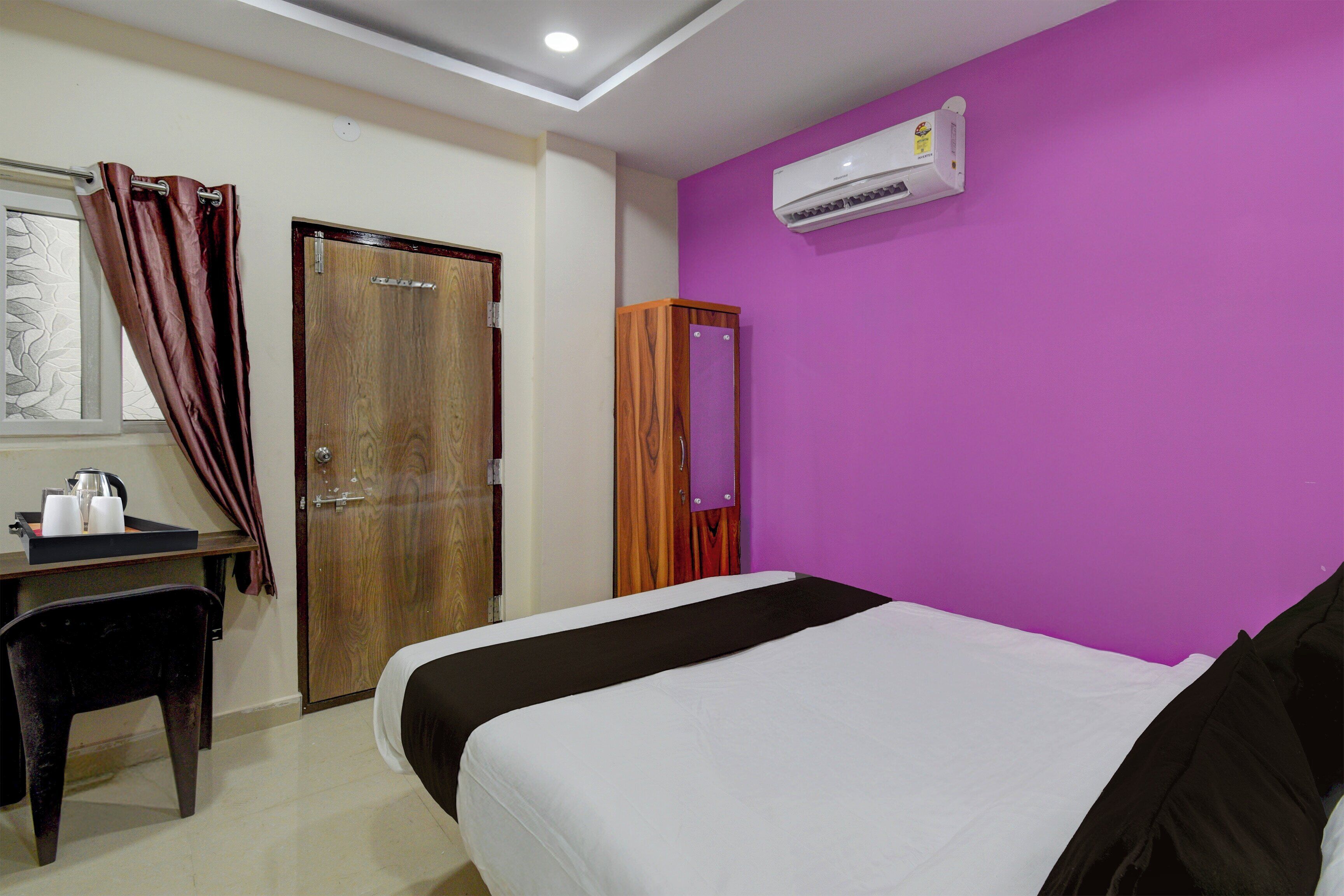 A hotel room with a bed, a desk with a chair, a wardrobe, and an air conditioning unit.