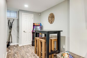 A room with a white door, a Pac-Man arcade game, a wooden table, and a woven dog bed. - Convenient, Cozy Home: Porches, Fire Pit, Office (Meriden)