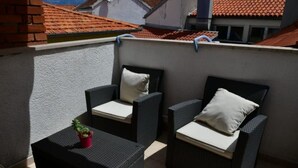 Ein Balkon mit zwei Korbstühlen, einem kleinen Tisch, einem Blumentopf und Blick auf ein rotes Ziegeldach.