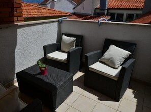 Ein Balkon mit zwei Korbstühlen, einem kleinen Tisch, einem Blumentopf und Blick auf ein rotes Ziegeldach.